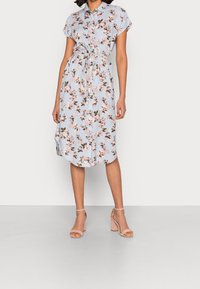 Donna che indossa un vestito floreale azzurro chiaro, lungo fino al ginocchio, con maniche corte, una cintura in vita e sandali beige con tacco a blocco.