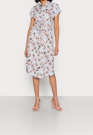 Frau in einem hellblauen, knielangen Blumenkleid mit kurzen Ärmeln, einem Taillengurt und beigefarbenen, Blockabsatzsandalen.
