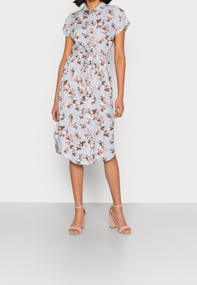 Donna che indossa un vestito floreale azzurro chiaro, lungo fino al ginocchio, con maniche corte, una cintura in vita e sandali beige con tacco a blocco.