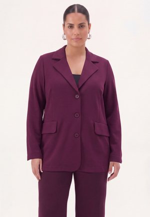 Vrouw met donker haar draagt een diep paarse, geknoopte blazer en bijpassende broek tegen een effen lichte achtergrond.