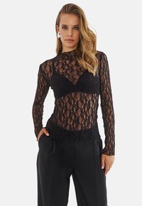 Czarna koronkowa bluzka z długim rękawem, z teksturowanym kwiatowym wzorem, przezroczystym materiałem i stójką, noszona na czarnym biustonoszu. Połączona z czarnymi spodniami.