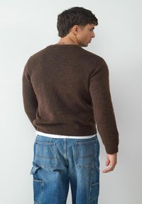 Brauner Strickpullover mit geripptem Saum und Bündchen, getragen über einem weißen Hemd, kombiniert mit blauen Jeans-Cargohosen mit mehreren Taschen.