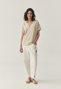 T-shirt beige à col en V avec des manches courtes, associé à un pantalon blanc avec des accents brodés. Le mannequin porte des sandales à talons plats beiges simples.