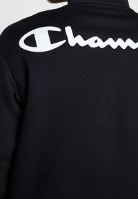 Felpa nera con una texture liscia, con un evidente logo bianco "Champion" sulla parte superiore della schiena. Design semplice e vestibilità comoda.
