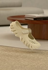 Chaussure Crocs beige avec des trous de ventilation et une semelle texturée flottant au-dessus d'un tapis beige, d'une table en bois et d'un canapé beige en arrière-plan.