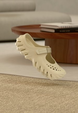 Scarpa Crocs beige con fori di ventilazione e suola strutturata che fluttua sopra un tappeto beige, un tavolo di legno e un divano beige sullo sfondo.