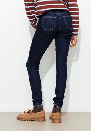 Personne portant un jean skinny bleu marine, une chemise à manches longues rayée marron, des chaussettes marron et des chaussures en suède marron clair à lacets, debout à l'intérieur.