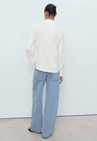 Cremefarvet bluse med lange ærmer og krave, parret med vide, lyseblå denimjeans. Glat tekstur, minimalistisk design og sideslids.