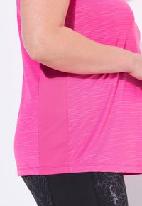 Primo piano di una persona che indossa una maglietta sportiva rosa e leggings neri a motivi, con il braccio appoggiato sul corpo.