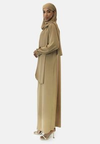 Vloeiende beige jurk met lange mouwen, losse pasvorm en zijzakken, gecombineerd met een bijpassende hijab en crèmekleurige sandalen met hoge hakken.