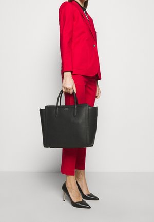 Femme en tailleur rouge tenant un grand sac à main en cuir noir, portant des escarpins noirs à bouts pointus, debout sur un fond blanc.