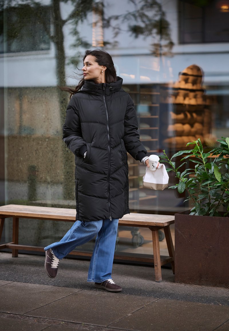 Schwarze gepolsterte Jacke mit Kapuze, Reißverschluss und Seitentaschen. Trägt sie über hellblauen Jeans und dunklen Sneakers, während sie eine Tasse Kaffee und einen Takeout-Beutel hält.