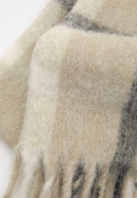 Uldhalstørklæde i beige og lys grå med en blød, fuzzy tekstur og frynser i enderne. Mønstret med et ternet design, som blander lyse og mørke nuancer.