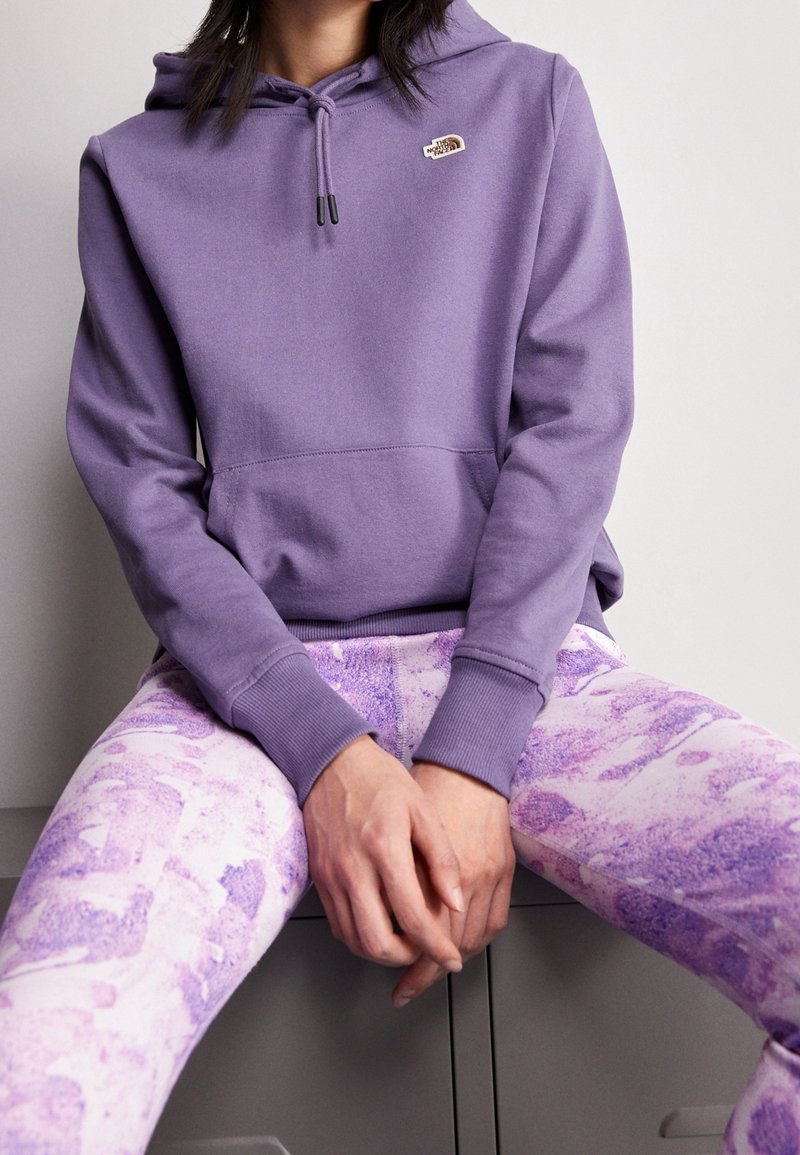 Personne assise portant un sweat à capuche violet avec un petit logo et un pantalon violet clair à motifs, mains jointes devant reposant sur les cuisses.