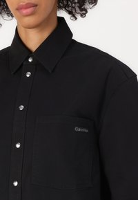 Camicia nera button-up in cotone con colletto a punta, bottoni argentati, taschino sul lato sinistro del petto e una piccola etichetta con logo sulla tasca.