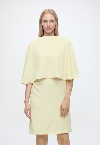Femme blonde vêtue d’une robe jaune pâle longueur genou avec un haut ample type cape, debout devant un fond blanc uni.