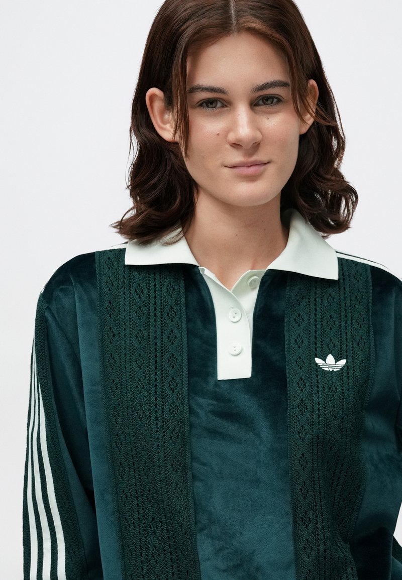 Green velvet polo shirt with a light green collar, three white buttons, and white embroidered logo. Features vertical lace pattern accents.