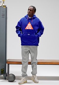 Μωβ φούτερ με πορτοκαλί τριγωνικό λογότυπο, γκρι φόρμα, και κρεμ αθλητικά παπούτσια. Το φόντο περιλαμβάνει ένα ντουλάπι και μια μπάλα μπάσκετ.