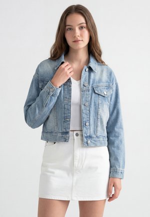 Ung kvinde med langt brunt hår iført en lyseblå denimjakke, hvid ribbet top og hvid nederdel, står foran en ensfarvet baggrund.