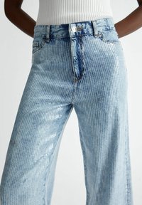 Vaaleansiniset farkut, joissa on leveät lahkeet, tekstuurinen ribbikuvio ja paljettikoristelu. Farkuissa on nappisuljenta ja etutaskut.