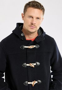 Homme avec les cheveux courts portant un manteau duffle bleu marine foncé avec des fermetures à bascule et une capuche, regardant droit devant lui avec une expression neutre.