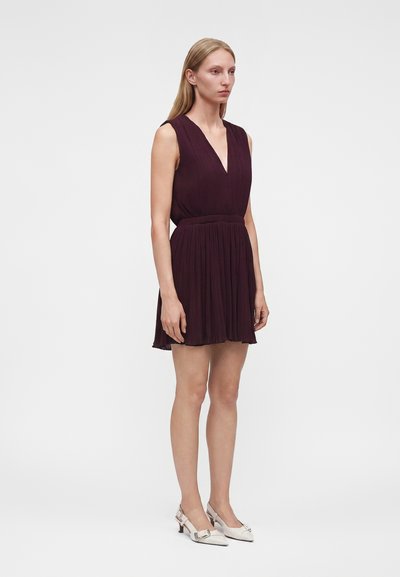 Femme en robe plissée sans manches de couleur violet foncé et chaussures blanches à talons bas, debout devant un fond clair uni.