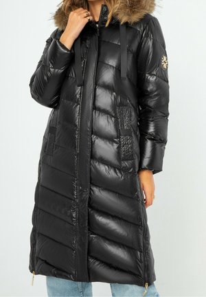 Long manteau d'hiver matelassé noir avec capuche bordée de fourrure, fermeture éclair sur le devant, poches latérales et écusson sur la manche.