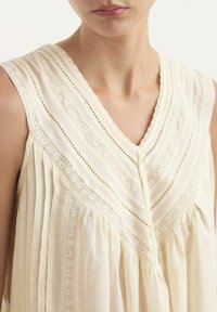 Blusa sin mangas de color crema claro con escote en V, que presenta pliegues, detalles de encaje y costuras sutiles. Tejido suave con un diseño fluido.