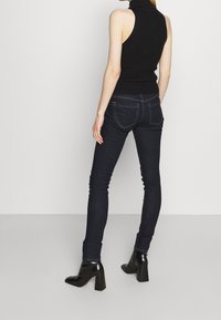 Top nero senza maniche con collo alto indossato con jeans skinny blu scuro, con vita alta, due tasche posteriori e stivaletti neri con tacco blocco.