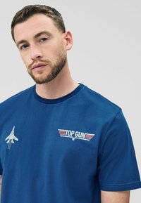 T-shirt in cotone blu con grafica di un aereo e logo "TOP GUN" in bianco e rosso, colletto rotondo e maniche corte.