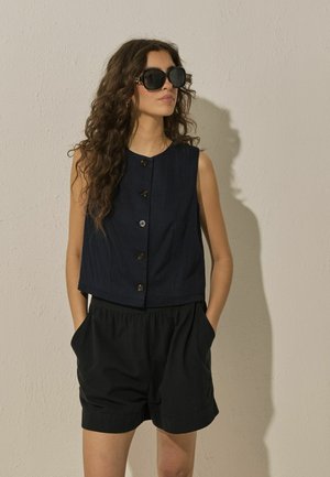 Femeie cu păr lung și creț, purtând ochelari mari de soare negri, un top bleumarin fără mâneci, cu nasturi, și pantaloni scurți negri, cu mâinile în buzunare.