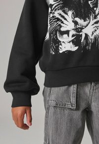 Sudadera negra con un gráfico de tigre blanco, con puños y dobladillo acanalados. Combinada con pantalones cargo grises, mostrando un detalle de bolsillo.