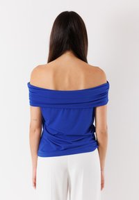 Top blu off-the-shoulder realizzato in tessuto morbido. Presenta dettagli arricciati lungo la scollatura e una vestibilità comoda. Abbinato a pantaloni bianchi.