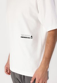 Tricou alb din bumbac, cu un eticheta grafică laterală în negru, ce conține text și un simbol de flacără. Mâneci scurte și o croială relaxată.