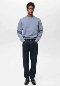 Felpa azzurra con polsini a coste, abbinata a pantaloni in denim blu scuro e scarpe nere. Il modello sta in piedi con le mani in tasca.