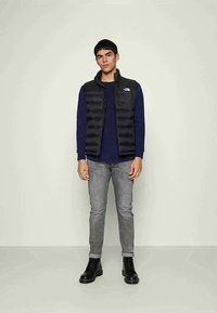 Ung mann står mot en ensfarget bakgrunn iført svart boblevest, marineblå langermet skjorte, grå jeans og svarte snøringstøvler.