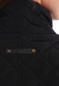 Veste matelassée noire avec un col haut et une étiquette en cuir portant le mot "LAUREN," fixée avec des boutons-pression en métal.