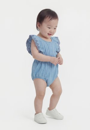 Peuter in een blauw perforatiepakje en witte sneakers, staand en glimlachend tegen een effen witte achtergrond.