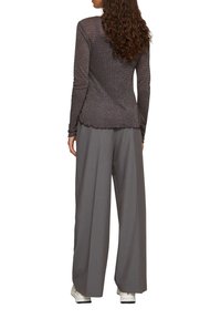 Langärmliges Oberteil aus dunklem Metallic-Stoff, an den Schultern tailliert mit einem gewellten Saum. Kombiniert mit lockeren grauen Hosen und weißen Schuhen.