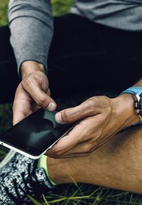 Czarny smartfon trzymany w dłoni z widocznymi palcami, na tle zielonej trawy. Smukły design z białą krawędzią. Widoczne swobodne ubranie i zegarek.