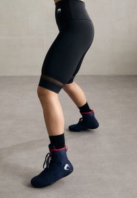Høye svarte sykkelshorts med mesh-paneler, parret med marineblå brytesko med røde detaljer og svarte såler, på et grått gulv.