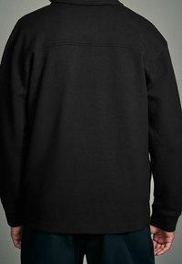 Sort strikket jakke med en tekstureret finish, der har en krave og synlige sømme. Bagfra ses lange ærmer og en lige kant.