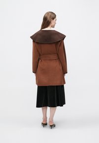 Palton maro din lână cu un guler mare și închis la culoare, talie cu curea și croială ajustată, purtat peste o fustă plisată neagră. Pantofi cu toc vizibili.