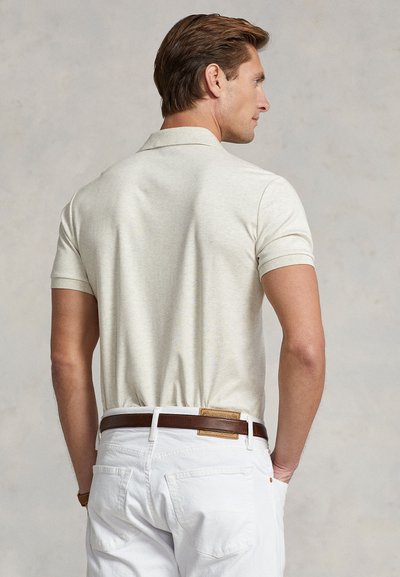 Mies, joka pukeutunut vaaleaan beigenväriseen lyhythihaiseen poolopaitaan ja valkoisiin housuihin, joissa on ruskea nahkavyö, seisoo sivuttain kädet taskuissa.