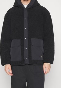 Schwarze Fleecejacke mit strukturierter Oberfläche, mit Kapuze, Druckknöpfen und zwei Fronttaschen mit Stoffakzenten.