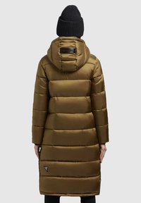 Personne portant un long manteau rembourré vert olive avec une capuche et un bonnet en tricot noir, se tenant de dos sur un fond clair.