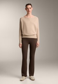 Bézs csíkos kötött pulóver széles, vízszintes nyakkivágással, barna harangszárú leggings-szel és világos színű sportcipőkkel párosítva. Finom anyag, amely enyhe textúrával rendelkezik.