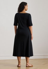 Czarna sukienka do kolan z rękawami za łokieć i okrągłym dekoltem. Do niej dopasowano beżowe sandały na paski na jasnej, drewnianej podłodze.