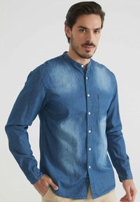 Homme aux cheveux et à la barbe bruns, portant une chemise en denim bleu à boutons avec un col mandarin, se tenant avec une main dans la poche de son pantalon beige.