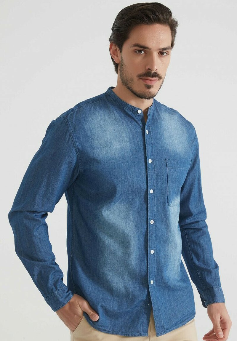 Homme aux cheveux et à la barbe bruns, portant une chemise en denim bleu à boutons avec un col mandarin, se tenant avec une main dans la poche de son pantalon beige.